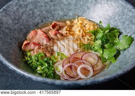 Vietnamese Cuisine - Beef Pho Noodle Soup or Pho Bo Soup. Served with Fresh Greens in Blue Bowl on Textured Dark Gray Table. Pho Bo Soup with Spicy Sauces