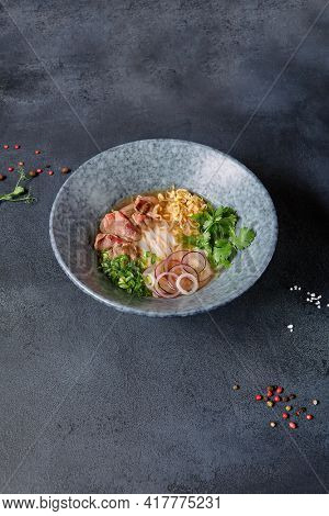 Vietnamese Cuisine - Beef Pho Noodle Soup or Pho Bo Soup. Served with Fresh Greens in Blue Bowl on Textured Dark Gray Table. Pho Bo Soup with Spicy Sauces