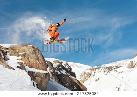 A professional skier makes a jump-drop from a high cliff against the blue sky leaving a trail of snow powder in the mountains. Photo from the slopes of Mount Elbrus. The concept of extreme sports and recreation in the mountains in winter. Copy the space. 