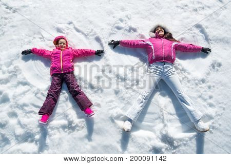 High angle view of cute daughter and mother playing and lying in the snow. Happy woman and smiling little girl having fun in snowdrift. Cheerful family making snow angel while lying on snow.