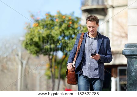 Young urban businessman professional on smartphone walking in street using mobile phone app texting sms message on smartphone wearing smart casual jacket. City lifestyle commute person walking.