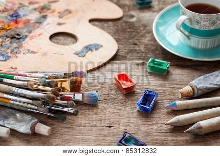 Paintbrushes, Artist Palette, Pencils, Coffee Cup, Watercolor And Oil Paints On Desk In Painter Stud