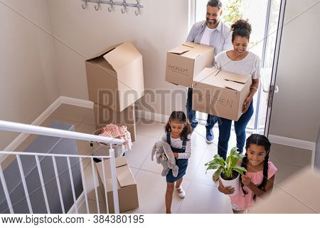 Ethnic family with two children carrying boxes and plant in new home on moving day. High angle view of happy smiling daughters helping mother and father with cardboard boxes in new house.