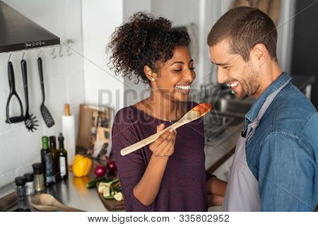 Young couple tasting tomato sauce while cooking in the kitchen. Cheerful man and smiling woman holding spatula in hand ready to taste red sauce. Multiethnic couple cooking together at home.