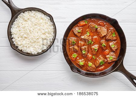 Goulash traditional Hungarian beef meat stew soup food cooked recipe with spicy gravy sauce in cast iron pan meal served with rice and chopped parsley on white wooden texture kitchen table background.