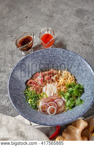 Vietnamese Cuisine - Beef Pho Noodle Soup or Pho Bo Soup. Served with Fresh Greens in Blue Bowl on Textured Dark Gray Table. Pho Bo Soup with Spicy Sauces