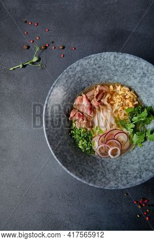 Vietnamese Cuisine - Beef Pho Noodle Soup or Pho Bo Soup. Served with Fresh Greens in Blue Bowl on Textured Dark Gray Table. Pho Bo Soup with Spicy Sauces. Top View