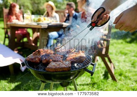 leisure, food, people and holidays concept - man cooking meat on barbecue grill for his friends at summer outdoor party