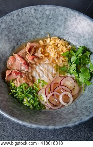 Vietnamese Cuisine - Beef Pho Noodle Soup or Pho Bo Soup. Served with Fresh Greens in Blue Bowl on Textured Dark Gray Table. Pho Bo Soup with Spicy Sauces