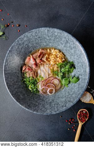 Vietnamese Cuisine - Beef Pho Noodle Soup or Pho Bo Soup. Served with Fresh Greens in Blue Bowl on Textured Dark Gray Table. Pho Bo Soup with Spicy Sauces. Top View