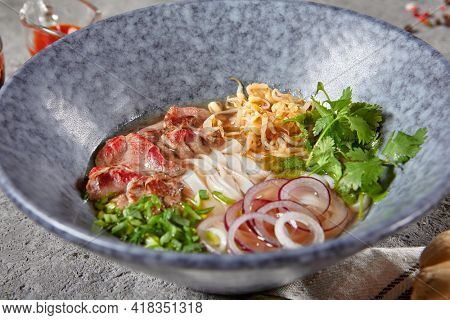 Vietnamese Cuisine - Beef Pho Noodle Soup or Pho Bo Soup. Served with Fresh Greens in Blue Bowl on Textured Dark Gray Table. Pho Bo Soup with Spicy Sauces