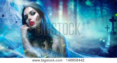 Beautiful Halloween Vampire Woman portrait. Beauty Sexy Vampire Vitch lady with blood on mouth posing in cementery, wearing spider web. Fashion Art design. Misterious Model girl with Halloween make up