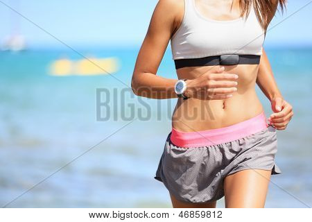 Runner woman with heart rate monitor running on beach with watch and sports bra top. Beautiful fit female fitness model training and working out outside in summer at part of healthy lifestyle.