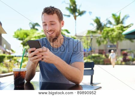 Man on cafe using smart phone app text messaging sms drinking iced coffee in summer. Handsome young casual man using smartphone smiling happy sitting outdoors. Urban male in his 20s.