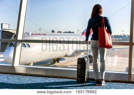 Travel tourist standing with luggage watching sunset at airport window. Unrecognizable woman looking at lounge looking at airplanes while waiting at boarding gate before departure. Travel lifestyle.